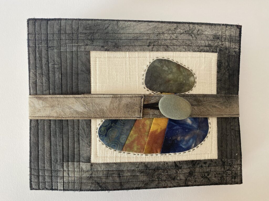 an image of a textile book made with rock blocks. the front cover shows a rock block surrounded with dark gray painted fabric and a closure with a real rock button