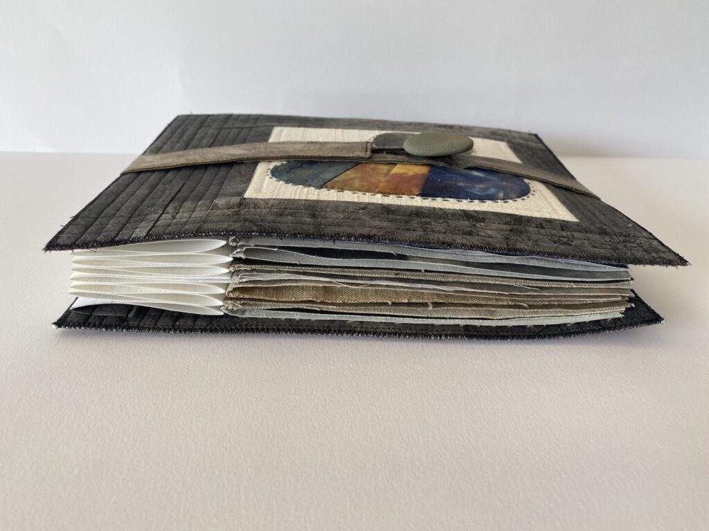 an image of a textile book made with rock blocks. the front cover shows a rock block surrounded with dark gray painted fabric and a closure with a real rock button. Seen from the side to show the spine and pages