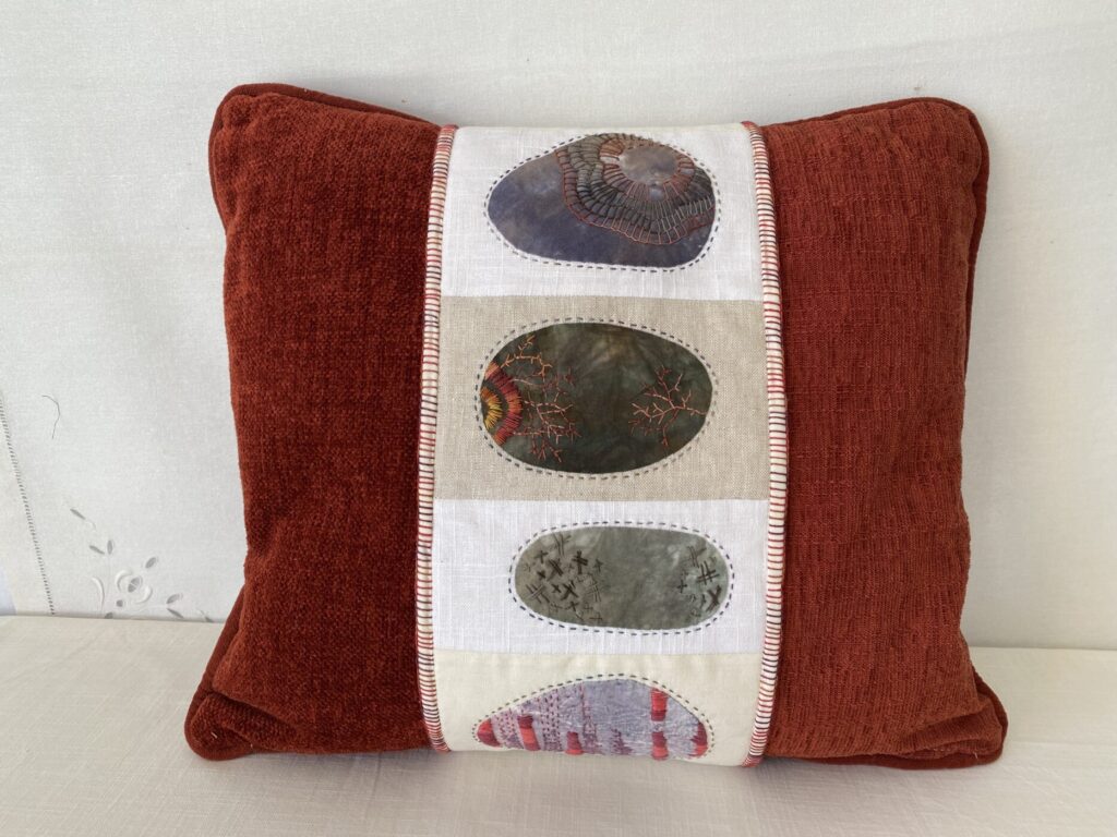 An image showing a pillow decorated with a band of rocks the pillow is burgundy red and the rocks are 4 in a column stitched together. Blanket stitch decorates the binding.