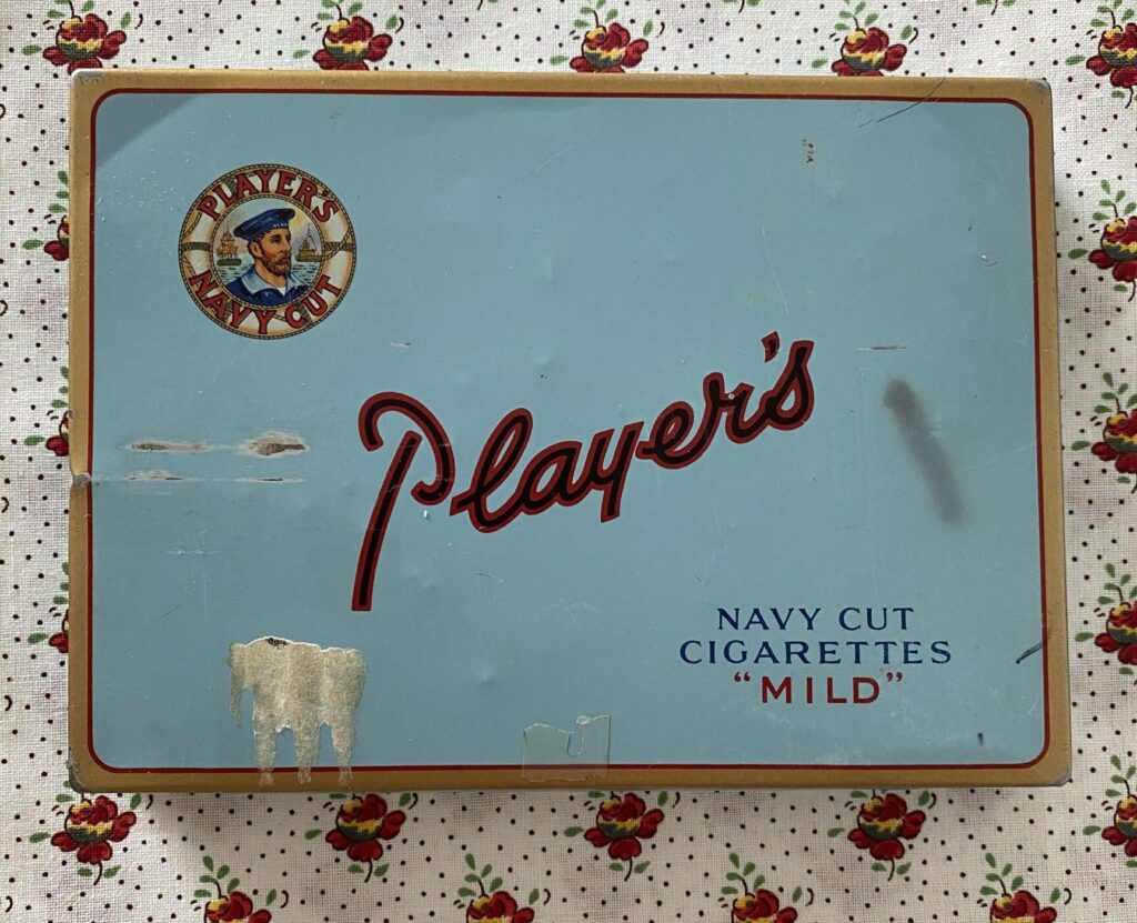 an image showing a Player's navy cut cigarettes blue tin on a piece of Provençal fabric with a white background, tiny red and yellow flowers and tiny dots