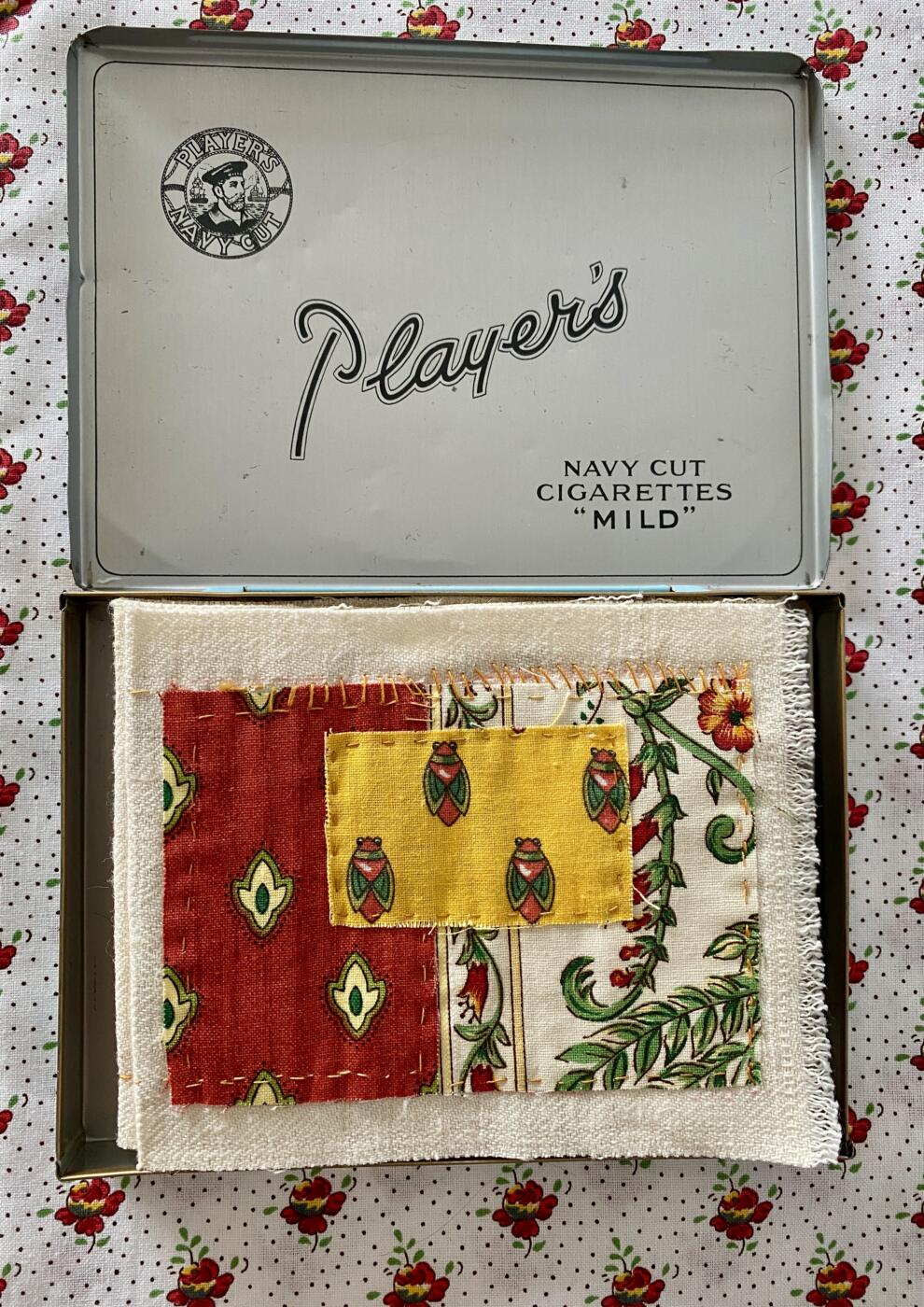 an image showing a Player's navy cut cigarettes tin on a piece of Provençal fabric with a white background, tiny red and yellow flowers and tiny dots. the box is open showing the tiny concertina book inside. The cover has three pieces of Provence fabric in reds, and yellows and white. The yellow piece has cicadas
