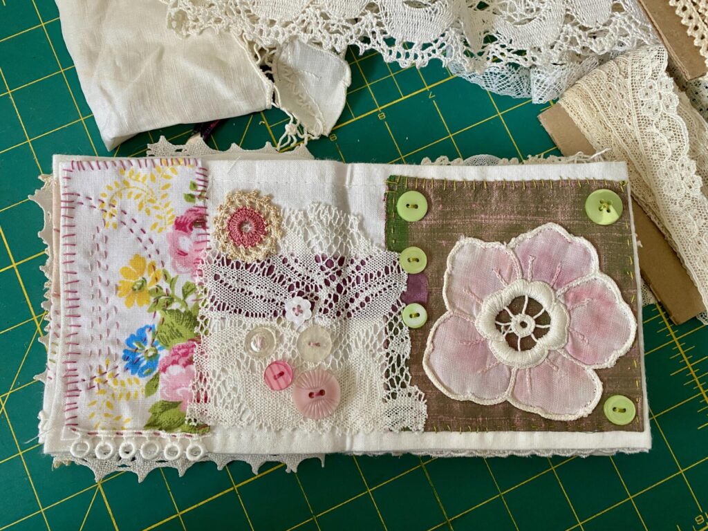 the image shows a two-page layout on the small concertina pink and green book. on the left, a piece of old vintage sheet with lace and buttons and on the right a piece of silk with a vintage lacey flower painted with pink pencils and a few green buttons