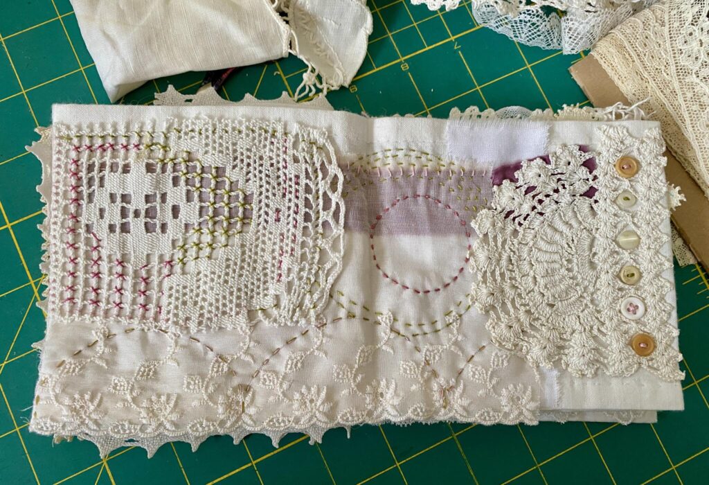 A two page layout on the small concertina book. this one is soft using a piece of vintage lacy fabric for the background over top of deep pink silk ribbon. a piece of doily on the left has stitching using cross stitches and on the right, a piece of vintage circular doily complements the design. round stitching in pink and green finish the piece
