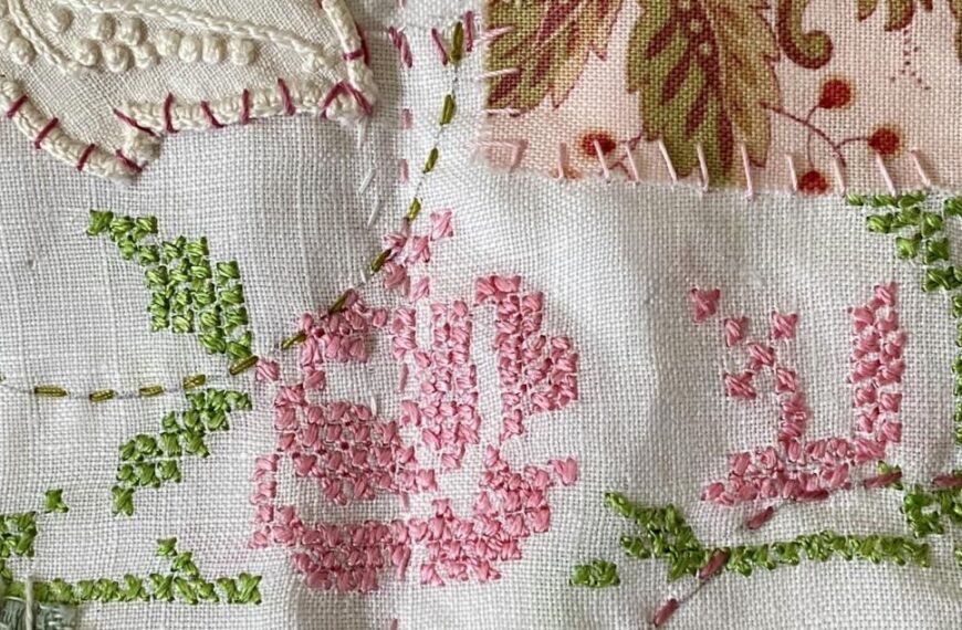 an image showing a detail from the first 2-page layout on the concertina book showing the small napkin with the cross stitched design in pink for the rose and green for the leaves. Plus some added fabrics and a few vintage buttons