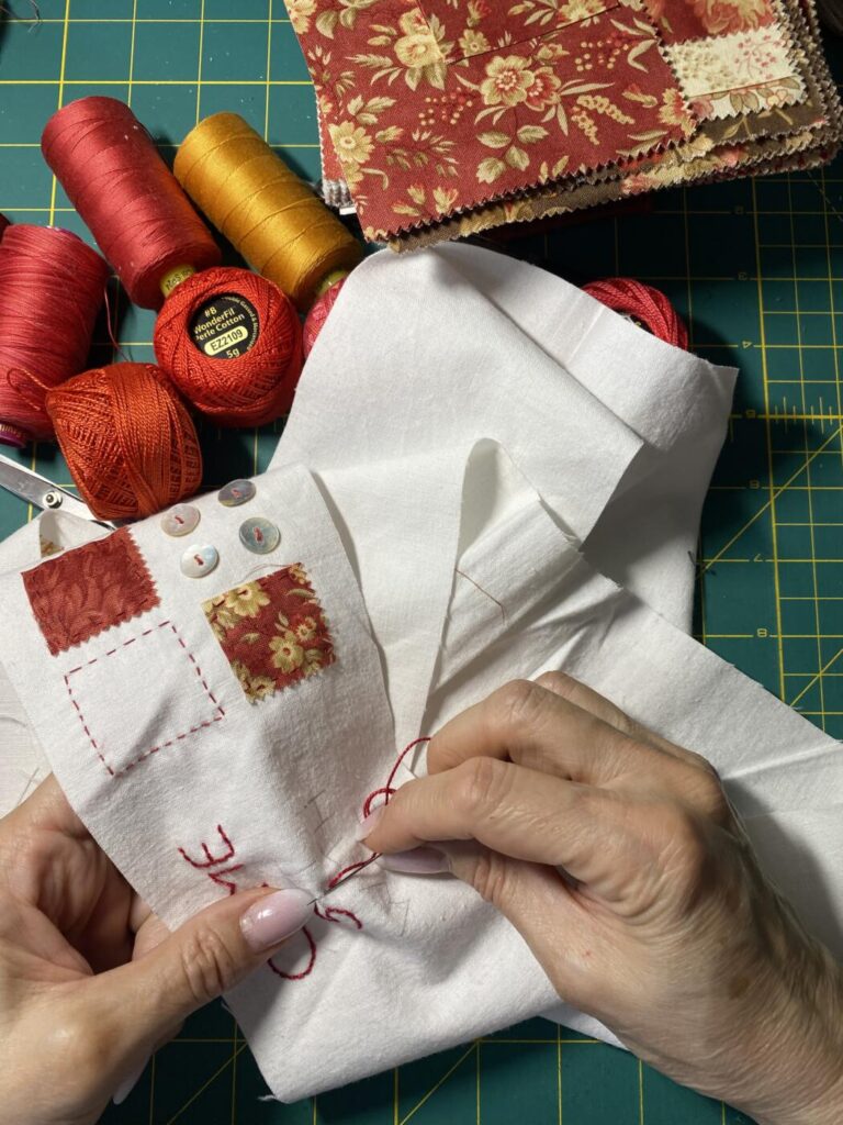an image showing two hands stitching letters on a strip of fabric. spools of threads and a stack of fabrics sit nearby